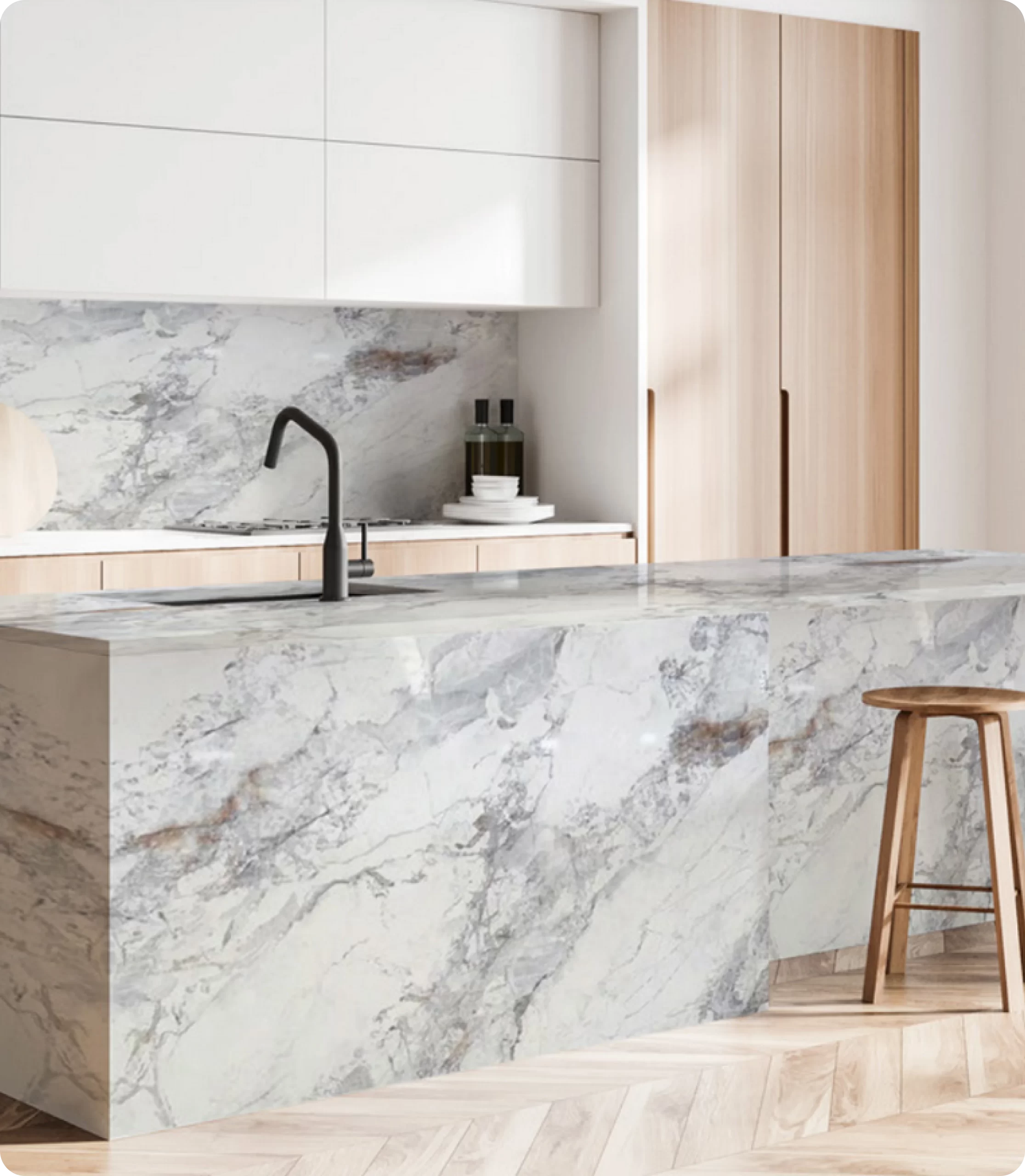 A modern kitchen by Slabmaster featuring white and wood cabinets, a marble countertop, and matching marble island with a black faucet and wooden bar stool. The light wood floor completes this stylish home space.