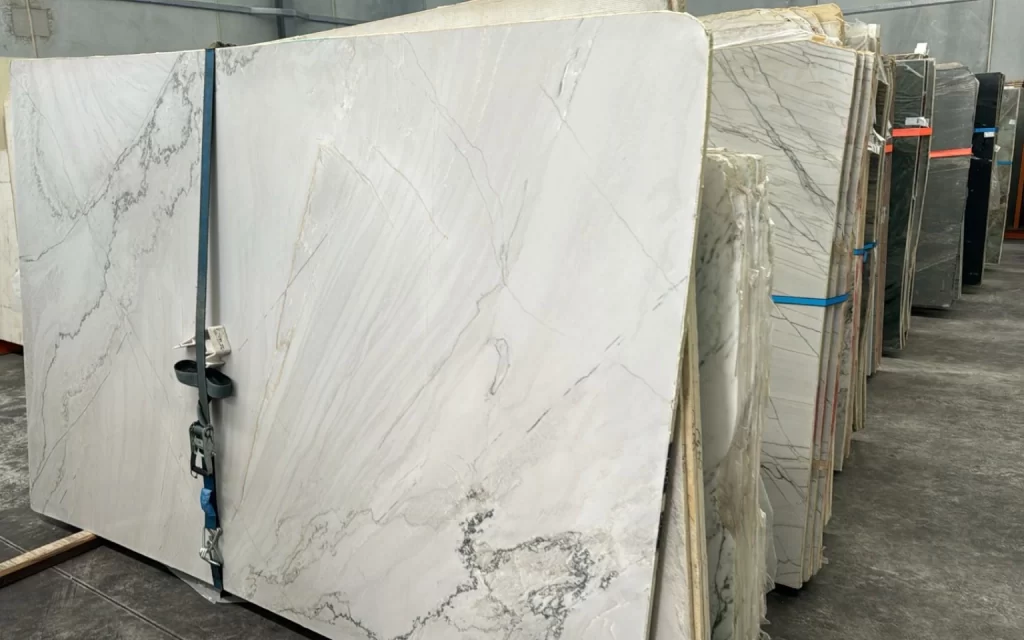 Large slabs of white marble with grey veining, each precisely cut to 20mm, are stacked upright in a warehouse setting, reminiscent of the timeless elegance of Santorini.