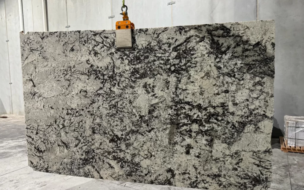 A large granite slab with gray and black patterns, reminiscent of the striking Branco White variety, is seen suspended by a metal clamp in an industrial setting.