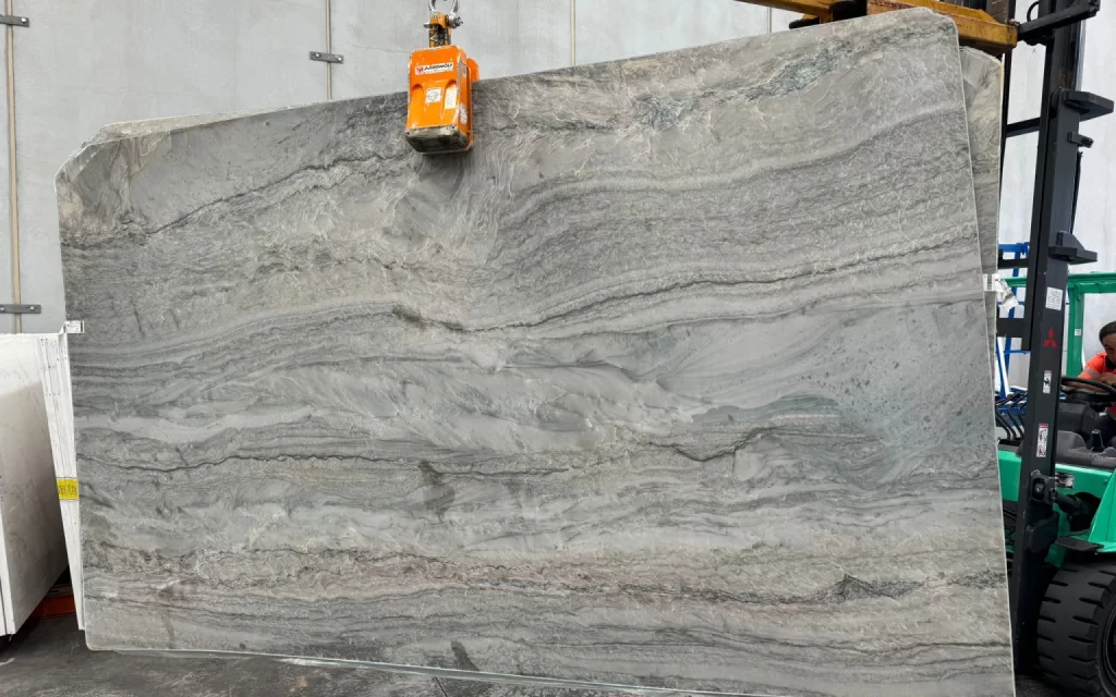 An elegant, large slab of grey marble with intricate veining patterns is being lifted by an orange crane inside a warehouse.