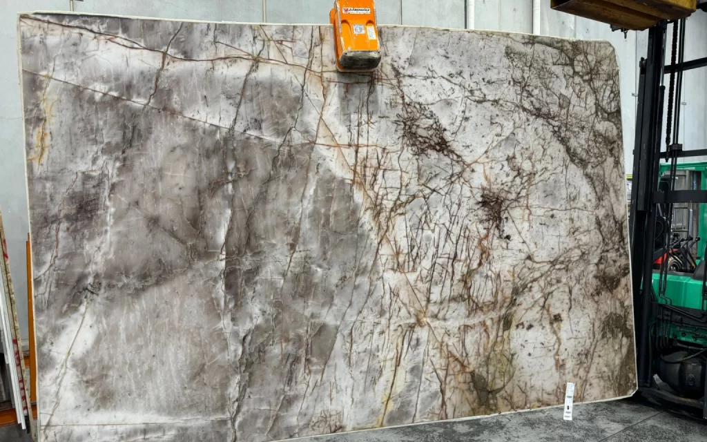 A large slab of stone with a mix of gray and brown veining patterns, reminiscent of an Ice Lake, is being moved using an orange lifting clamp attached to a forklift in an industrial setting.