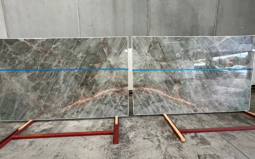 Two large Jadore slabs of polished stone with green and pinkish veins are displayed on wooden support frames against a gray wall in a warehouse.