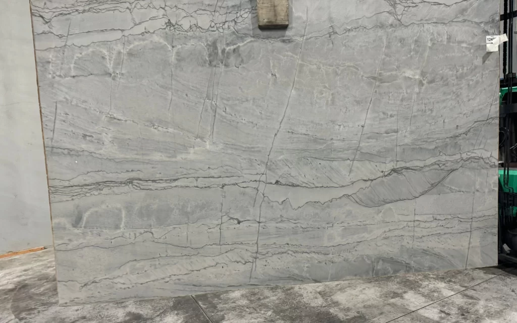 A close-up photo of a large, light gray marble slab with natural veining patterns reminiscent of the tranquil waters around the Maldives. The rectangular slab's visible edges emphasize its sturdy presence as it stands upright on a concrete floor.