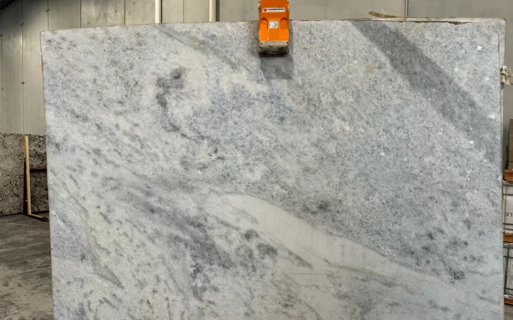 A large, gray marble slab with white and dark gray veining, reminiscent of marmore, is held upright by an orange lifting clamp in an industrial setting.