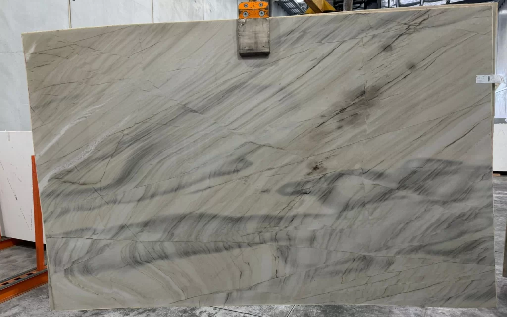 A large slab of polished marble from the Naica mines, featuring exquisite gray and white veining patterns, is held upright by a mechanical clamp in an indoor industrial setting.