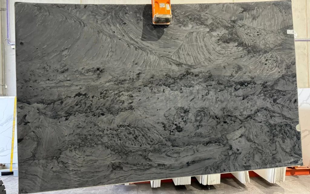 A large slab of grey marble with intricate swirling patterns is displayed in an indoor setting, held upright by sturdy equipment.