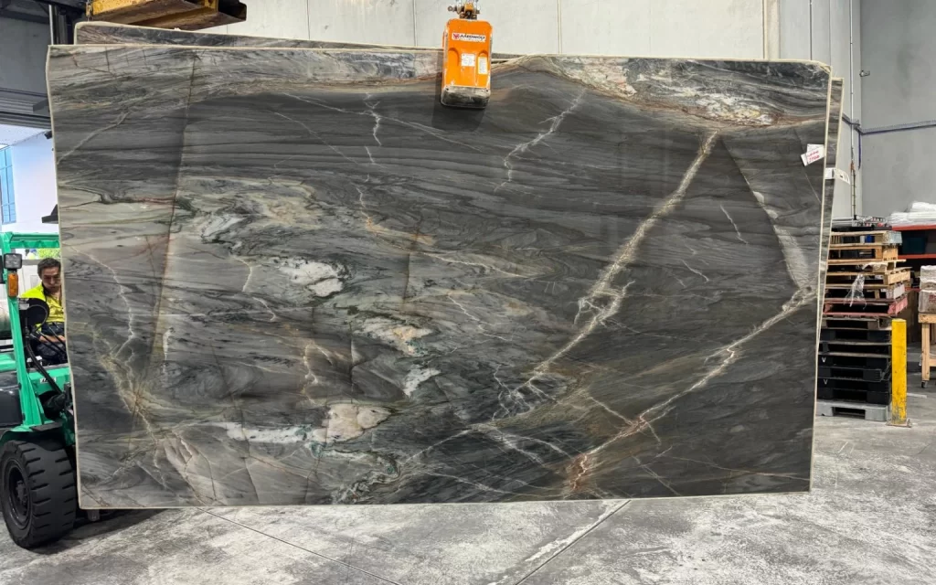 A large slab of dark marble with white and gray veining, reminiscent of the ocean's depths, is suspended by a crane in an industrial setting. A forklift and warehouse shelves are visible in the background, casting a stark contrast to the marble's natural elegance.