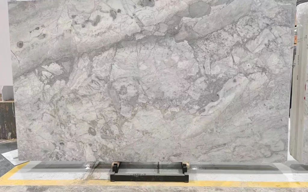 A large slab of Super Grey marble with intricate veining patterns, displayed upright on a supporting stand in a showroom or warehouse.