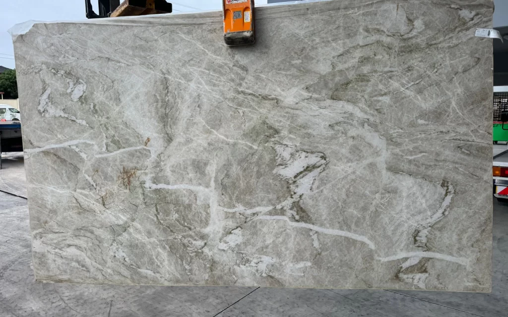 A large slab of light gray and white marble with a polished surface is being lifted by an orange clamp. The slab, reminiscent of the exquisite marble seen in the Taj Mahal, has prominent veining and is situated in an outdoor area, echoing destinations frequented by avid travel enthusiasts.