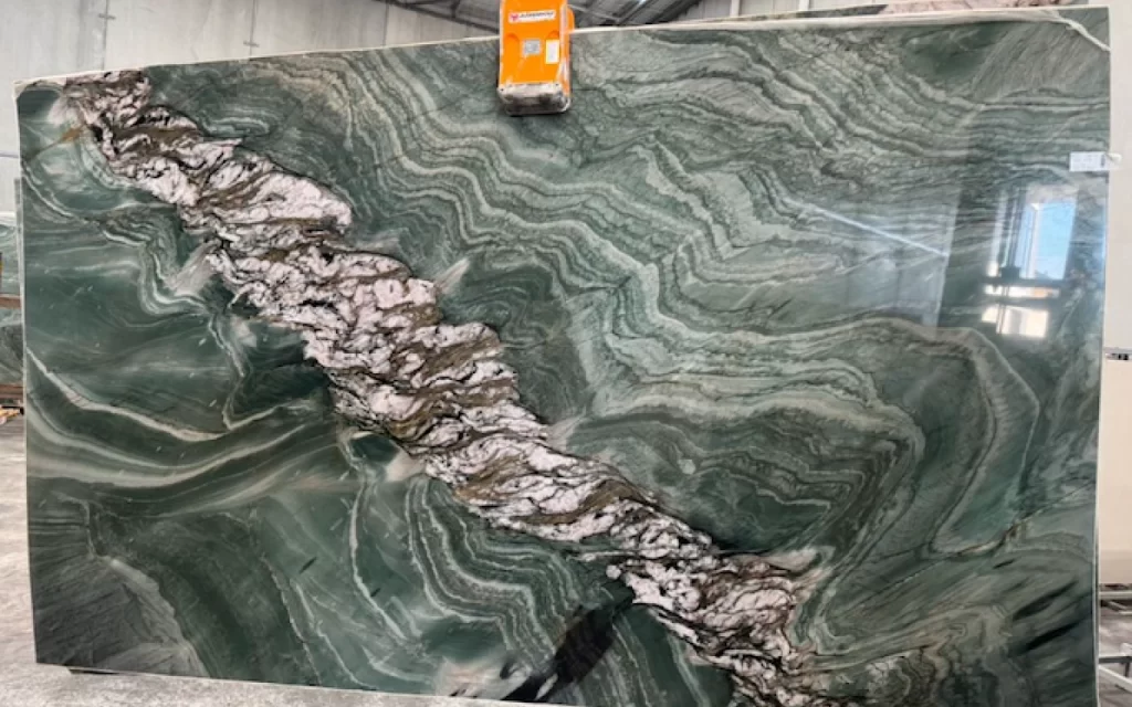 A large, polished green marble slab with a prominent white and gray vein running diagonally through the center, displayed vertically in an industrial setting, evokes tropical elegance amidst its urban surroundings.