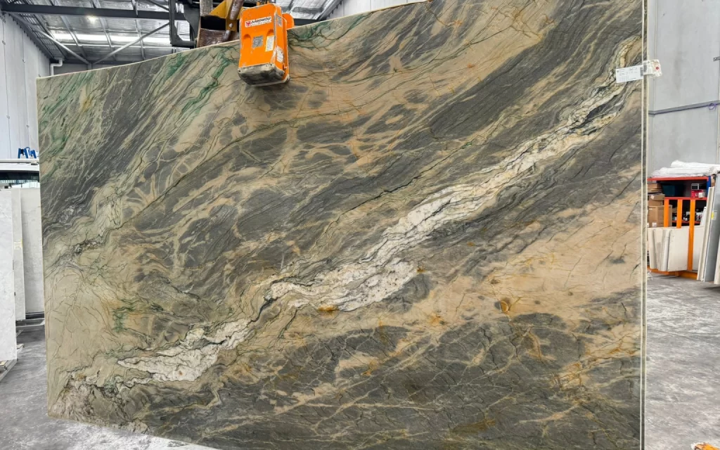 A large slab of natural stone, reminiscent of Van Gogh paintings with its mix of green, gray, and beige patterns, is held by an overhead lifting device inside an industrial warehouse.