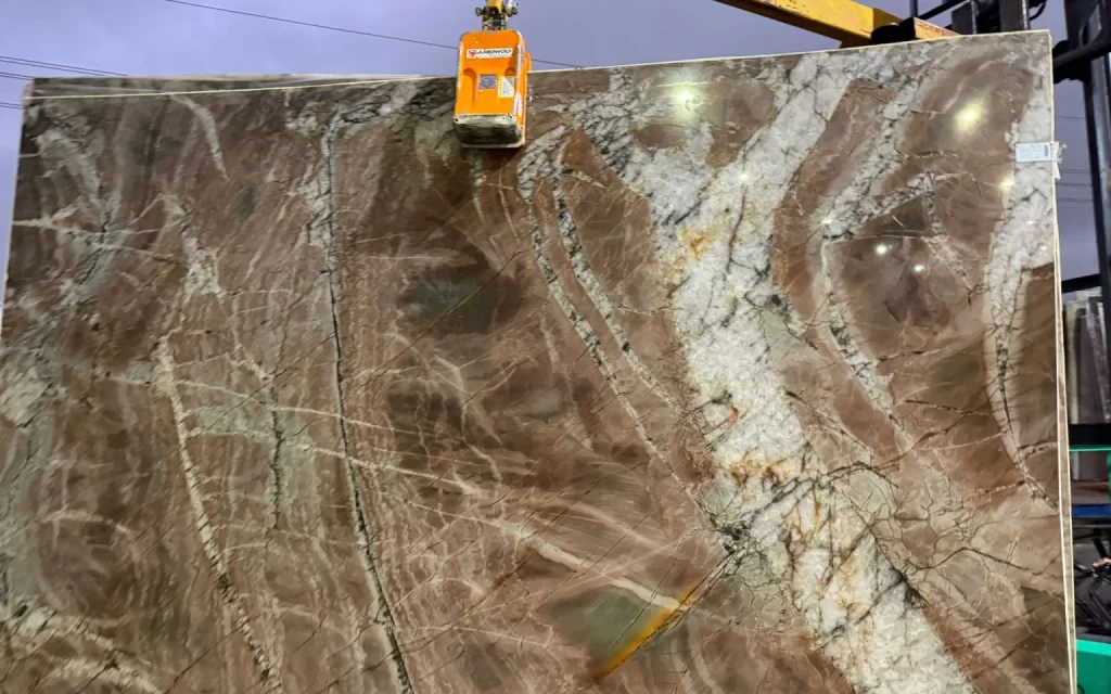 A large slab of brown and beige marble with intricate veining patterns, reminiscent of Volupia, is being lifted by a crane.