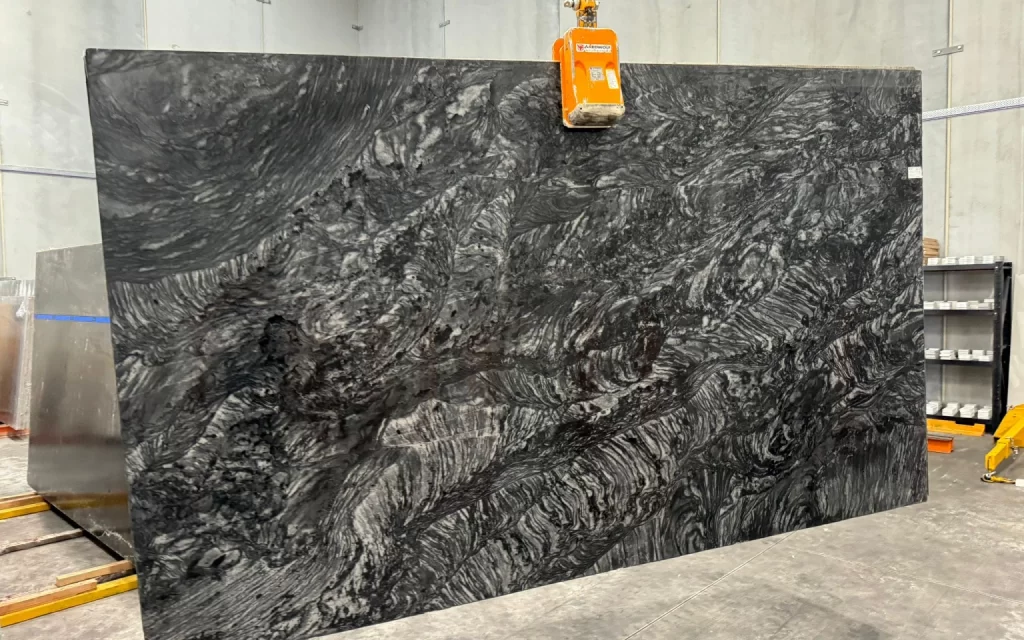 A large slab of grey-hued marble with white and gray veins is being held upright by a mechanical clamp in a warehouse. Shelves and other equipment are visible in the background, lending an industrial feel to this scene.