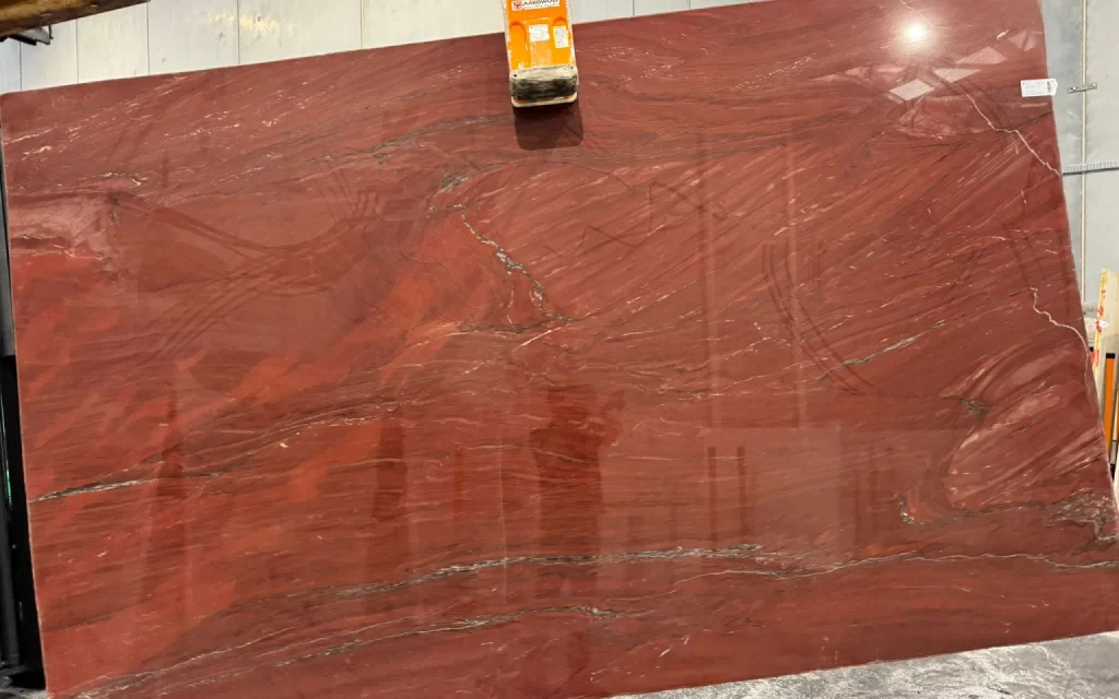 A large Xanga-red marble slab with intricate natural patterns is held upright by a clamp in a warehouse setting.