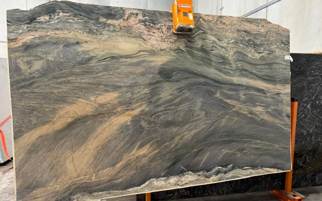 A large, multicolored marble slab with shades of gray, beige, and black stands upright in a workshop setting, held by an orange clamp and accented with subtle streaks of amarelo.