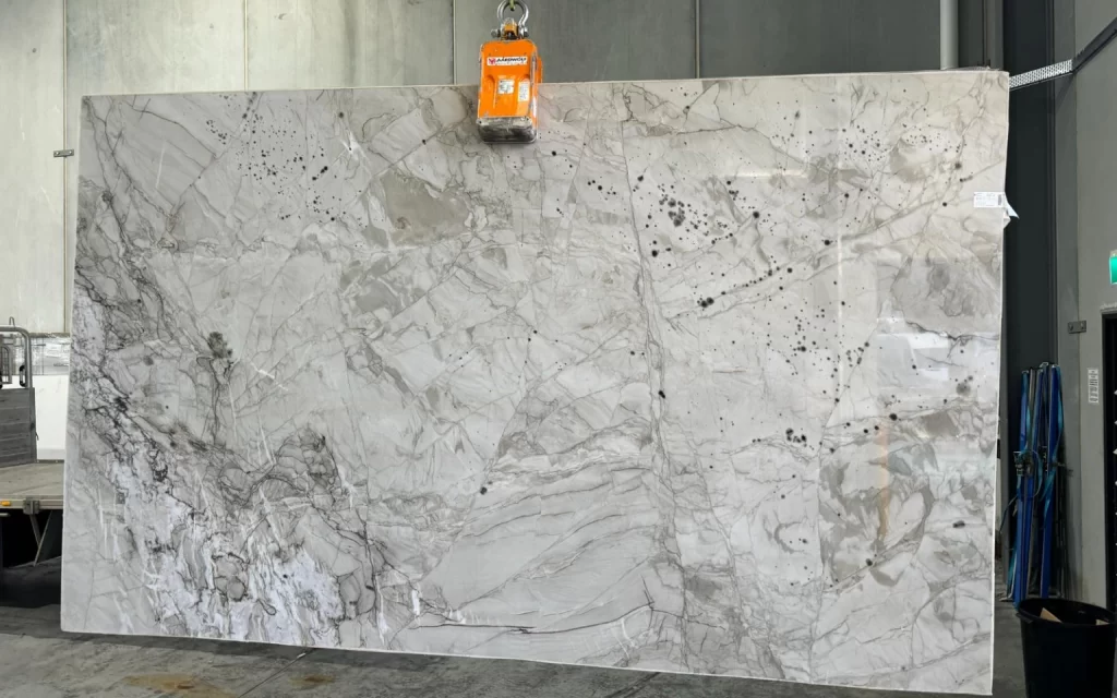 A large Apolo White marble slab is suspended by an orange lifting device in an industrial setting. The marble displays grey and white veining with striking black spots.