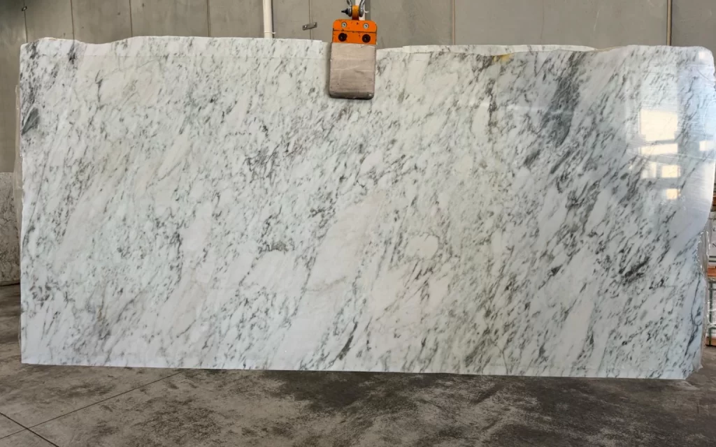 A large white and gray marble slab, reminiscent of the ethereal Aurora, is lifted by a clamp in an industrial setting with concrete floors and walls.