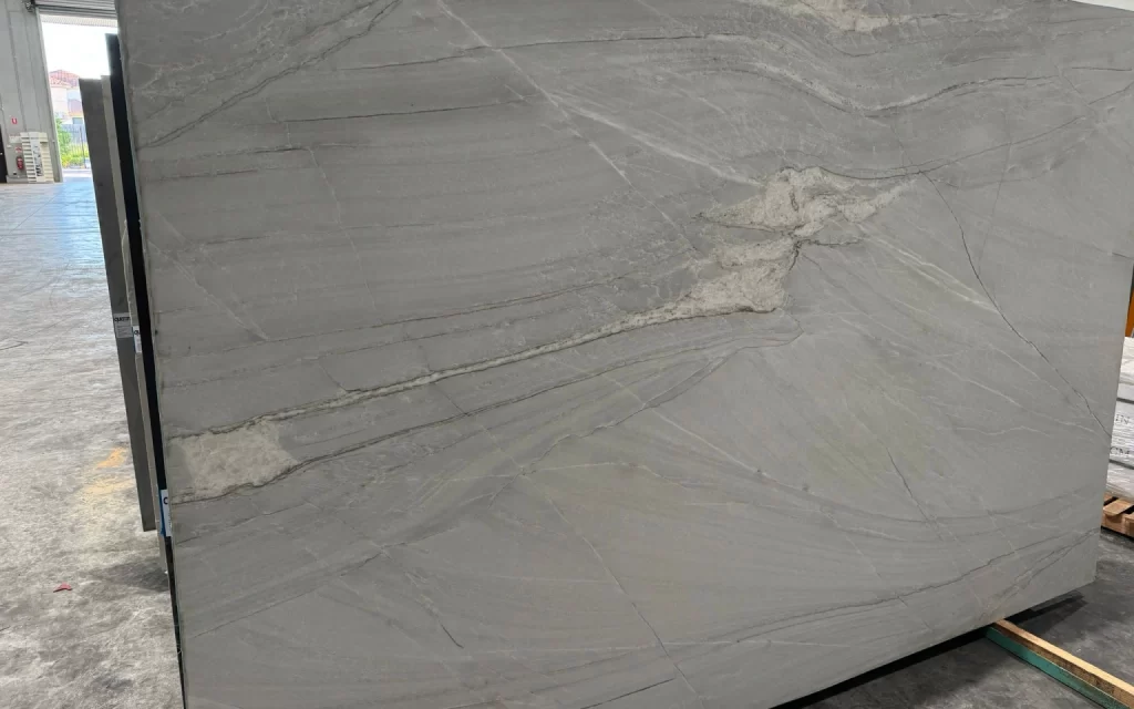 A large slab of gray stone with natural veins and patterns, known as Black Storm, stands in a warehouse. The slab has a polished surface, with the background showing an industrial area.