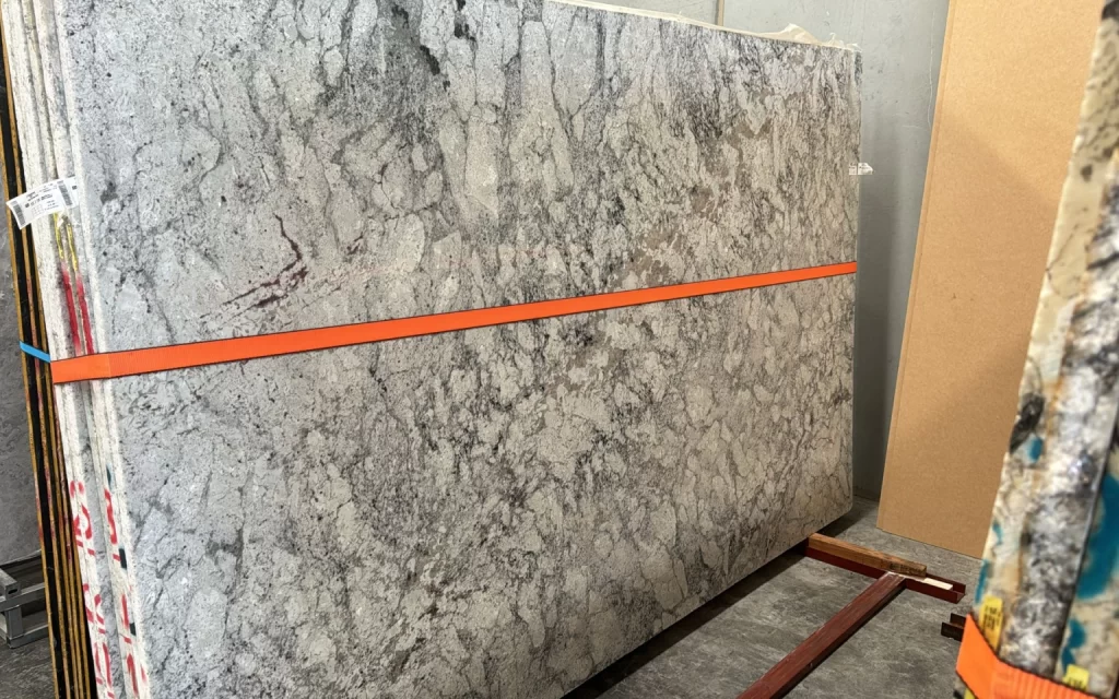 A large slab of Romano granite with a predominantly grey and white marbled pattern, secured with an orange strap, stands upright in a storage area next to other slabs and materials.