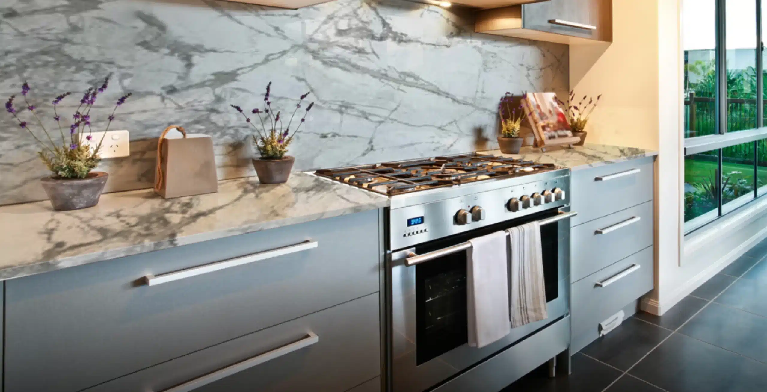 Modern kitchen with a stainless steel stove, marble backsplash, and light gray cabinets. Small potted plants and decorative items are on the countertop. Large window on the right.