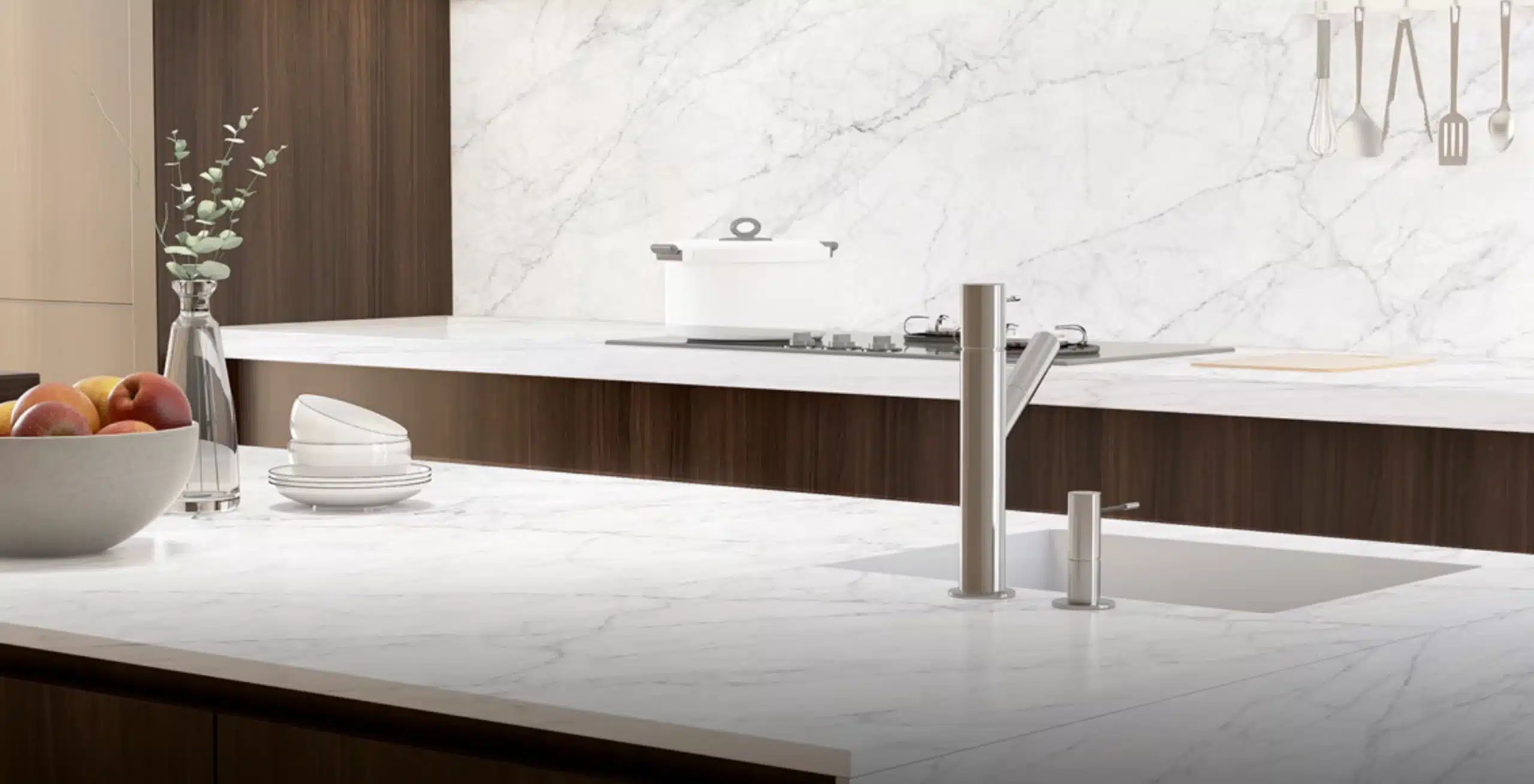 Modern kitchen with a marble countertop and backsplash. A sink with a faucet is in the foreground, and a stovetop with a white pot is in the background. A bowl of apples and stacked plates are on the counter.