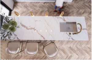 Overhead view of a Belizza d’Oro marble kitchen island with built-in sink, gold faucet, three beige chairs, pendant lights, and plant decor; person walking by in the background.
